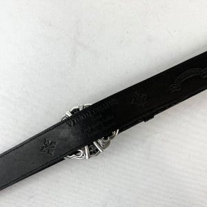 K&ouml;nnte beinhalten: Schwarzer Lederg&uuml;rtel mit silberner Schnalle. Die Schnalle hat ein dekoratives Design mit schwarzen Akzenten. Der G&uuml;rtel ist mit floralen Mustern und dem Markennamen "Vigor Deville" gepr&auml;gt.