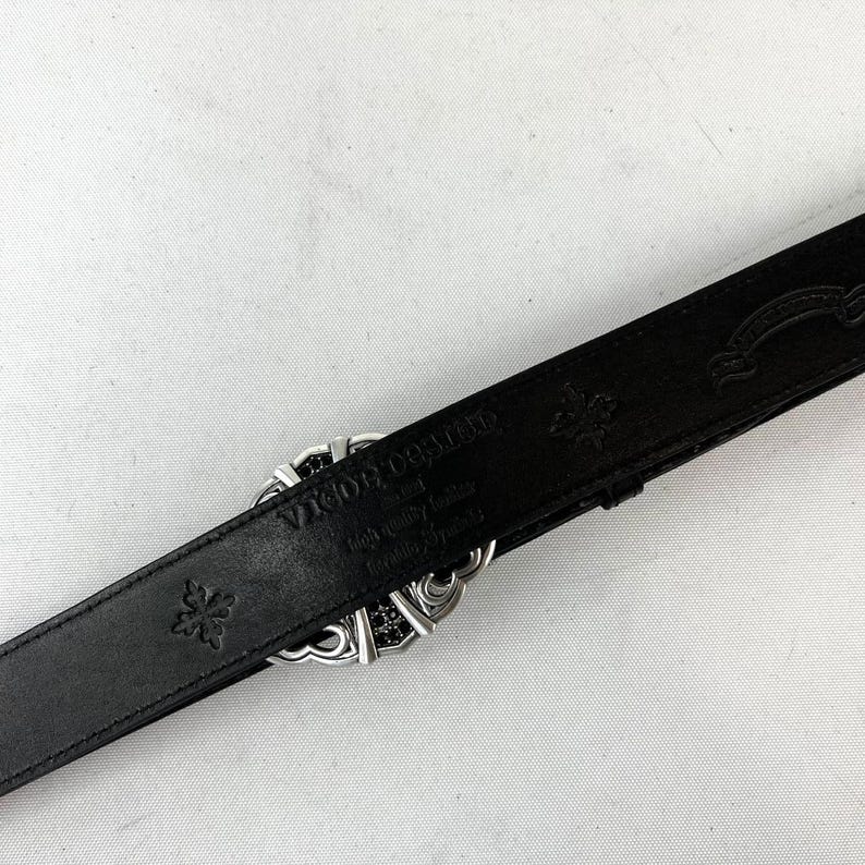 K&ouml;nnte beinhalten: Schwarzer Lederg&uuml;rtel mit silberner Schnalle. Die Schnalle hat ein dekoratives Design mit schwarzen Akzenten. Der G&uuml;rtel ist mit floralen Mustern und dem Markennamen "Vigor Deville" gepr&auml;gt.