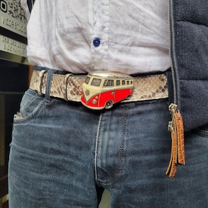 May include: A belt with a snakeskin pattern and a red and cream-colored Volkswagen bus buckle. The belt is worn with blue jeans and a white shirt. The buckle is made of metal.