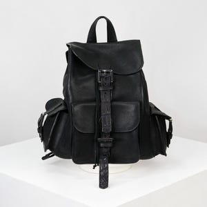 Può includere: Zaino in pelle nera con chiusura a patta, due tasche frontali e tasche laterali. Lo zaino ha un manico superiore e spallacci regolabili. La cinghia di chiusura con fibbia ha una trama a coccodrillo. La borsa è appoggiata su una superficie bianca.