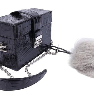 Puede incluir: Un bolso negro rectangular con un patrón texturizado similar al de un cocodrilo. Cuenta con una correa y cierre de cadena plateada, un asa superior y un accesorio gris y esponjoso.