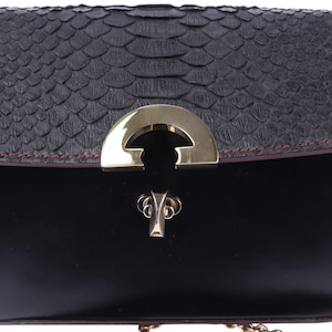 May include: Black handbag with a textured, dark grey flap and a smooth, black body. The flap has a scale-like pattern and is trimmed with red stitching. A gold-coloured clasp secures the bag, which has a gold chain strap.