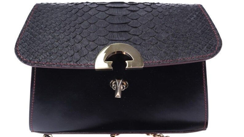 May include: Black handbag with a textured, dark grey flap and a smooth, black body. The flap has a scale-like pattern and is trimmed with red stitching. A gold-coloured clasp secures the bag, which has a gold chain strap.