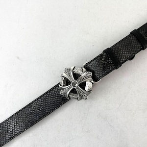 Może przedstawiać: Czarny skórzany pasek ze srebrną, ozdobną klamrą. Klamra ma wzór krzyża z osadzonymi kamieniami szlachetnymi. Pasek ma fakturę skóry węża.