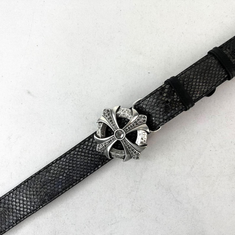 May include: A black leather belt with a silver-coloured, ornate buckle. The buckle features a cross design with embedded gemstones. The belt has a snakeskin texture.