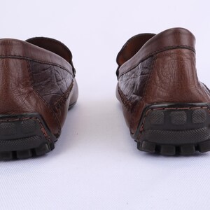 May include: A pair of brown leather loafers with a textured finish. The shoes have a rounded toe and a rubber sole. The image is a rear view of the shoes, showcasing the heel and sole details.