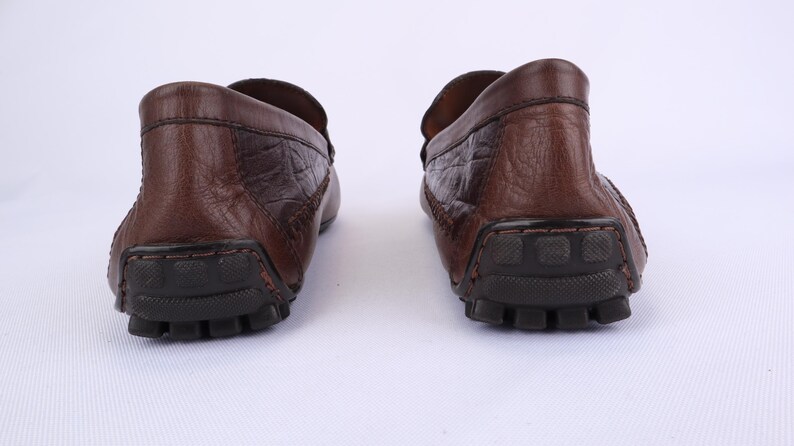 May include: A pair of brown leather loafers with a textured finish. The shoes have a rounded toe and a rubber sole. The image is a rear view of the shoes, showcasing the heel and sole details.