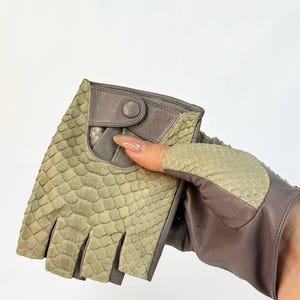 Puede incluir: Guantes sin dedos con textura de reptil en tonos verdes y grises. Los guantes tienen cierre de botón y están hechos de una combinación de materiales. El diseño presenta un patrón de escamas único.