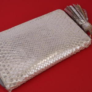 May include: A silver-colored, rectangular wallet with a textured, snakeskin-like pattern. The wallet has a zipper closure and a decorative tassel. The background is a solid red color.