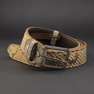 May include: A belt featuring a textured, snakeskin-patterned surface in shades of beige and brown. The belt includes a rectangular, dark metallic buckle and a matching snakeskin-patterned loop near the buckle, with a dark brown interior lining.