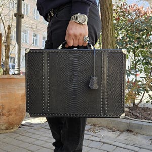 May include: A black briefcase with a snakeskin pattern, silver rivets, and metal corner protectors. The briefcase has a handle and a matching leather tag. The case is rectangular and appears to be made of leather.