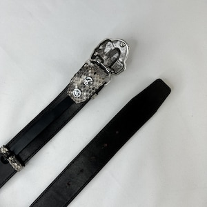Pode incluir: Cinto de couro preto com fivela prateada. A fivela tem um design decorativo e &eacute; presa a uma se&ccedil;&atilde;o do cinto com um padr&atilde;o de pele de cobra. O cinto est&aacute; desabotoado e repousa sobre uma superf&iacute;cie branca.