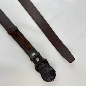 May include: A brown leather belt with a decorative buckle. The belt has a dark brown color and a detailed buckle design. The buckle is a dark metal color with two silver-colored studs. The belt is approximately 1.5 inches wide.