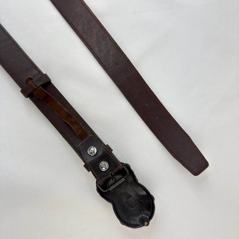 May include: A brown leather belt with a decorative buckle. The belt has a dark brown color and a detailed buckle design. The buckle is a dark metal color with two silver-colored studs. The belt is approximately 1.5 inches wide.