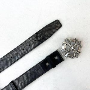 May include: A black belt with a snakeskin pattern and a silver cross buckle. The buckle has a hammered texture and is embellished with small, dark stones. The belt is made of leather.
