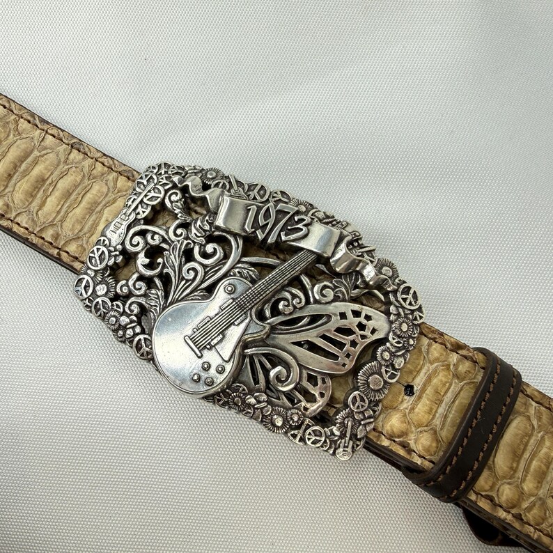 K&ouml;nnte beinhalten: Silberfarbene G&uuml;rtelschnalle mit Gitarren- und Schmetterlingsdesign, mit dem Jahr "1973" auf einem Banner. Der G&uuml;rtel hat ein beiges Schlangenmuster mit braunem Lederrand.