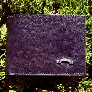 Può includere: Un portafoglio bifold viola scuro con una finitura in pelle di struzzo testurizzata dal caratteristico motivo a pori. Un piccolo emblema metallico argentato e curvo è fissato nell'angolo in basso a destra con il testo inciso "Since 1989".