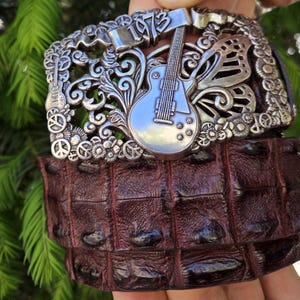 May include: A silver-colored belt buckle with a guitar and butterfly design, featuring the year "1973". The belt is made of dark brown, textured leather.