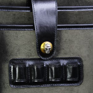 May include: Close-up of a dark green bag with black leather accents. The bag features a black leather strap with a gold skull button closure. Below the strap is a black leather holder with four compartments.