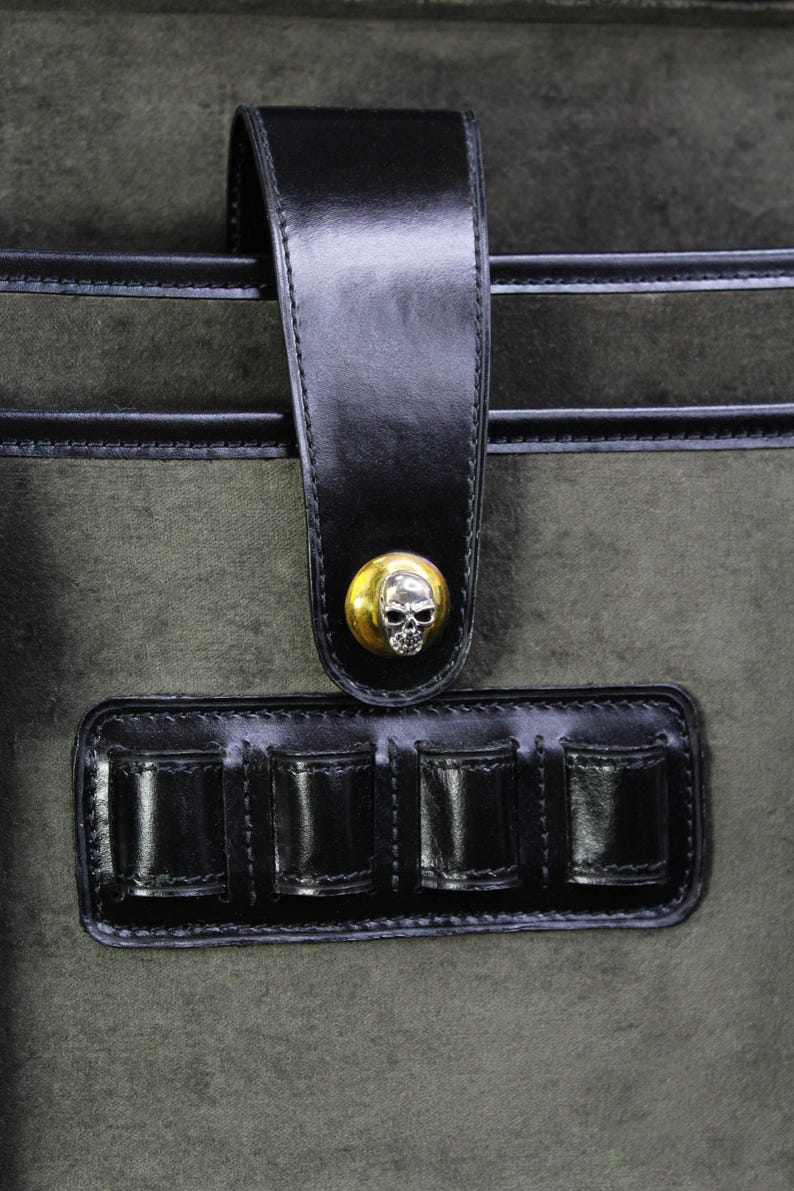 May include: Close-up of a dark green bag with black leather accents. The bag features a black leather strap with a gold skull button closure. Below the strap is a black leather holder with four compartments.