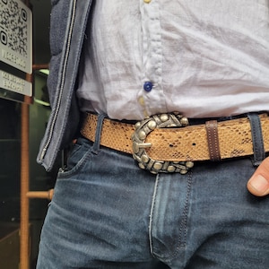 May include: A tan snakeskin-effect belt with a large silver buckle. The belt is worn with blue jeans and a white button-down shirt. The buckle has a decorative design with small silver studs.