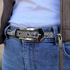 Könnte beinhalten: Ein Gürtel mit Schlangenmuster und einer silberfarbenen Schnalle wird mit einer Jeans getragen. Der Gürtel besteht aus einem hellgrauen und braunen Material. Die Schnalle hat eine rechteckige Form.