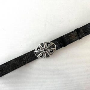 Op de afbeelding: Zwarte leren riem met een zilverkleurige ovale gesp. De gesp heeft een decoratief ontwerp met zwarte accenten. De riem heeft een textuurpatroon.