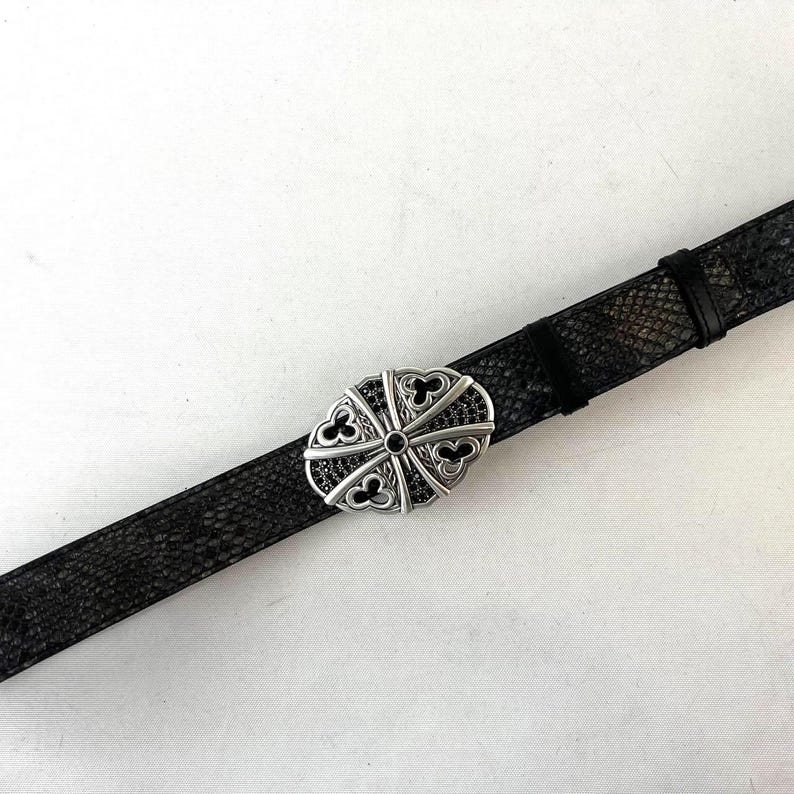 K&ouml;nnte beinhalten: Schwarzer Lederg&uuml;rtel mit einer silberfarbenen ovalen Schnalle. Die Schnalle hat ein dekoratives Design mit schwarzen Akzenten. Der G&uuml;rtel hat ein strukturiertes Muster.