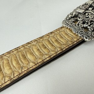 K&ouml;nnte beinhalten: Nahaufnahme eines beigefarbenen Lederg&uuml;rtels mit Schlangenmuster. Der G&uuml;rtel hat eine dekorative Silberschnalle mit aufwendigen Designs. Der G&uuml;rtel ist mit braunem Faden an den Kanten gen&auml;ht.