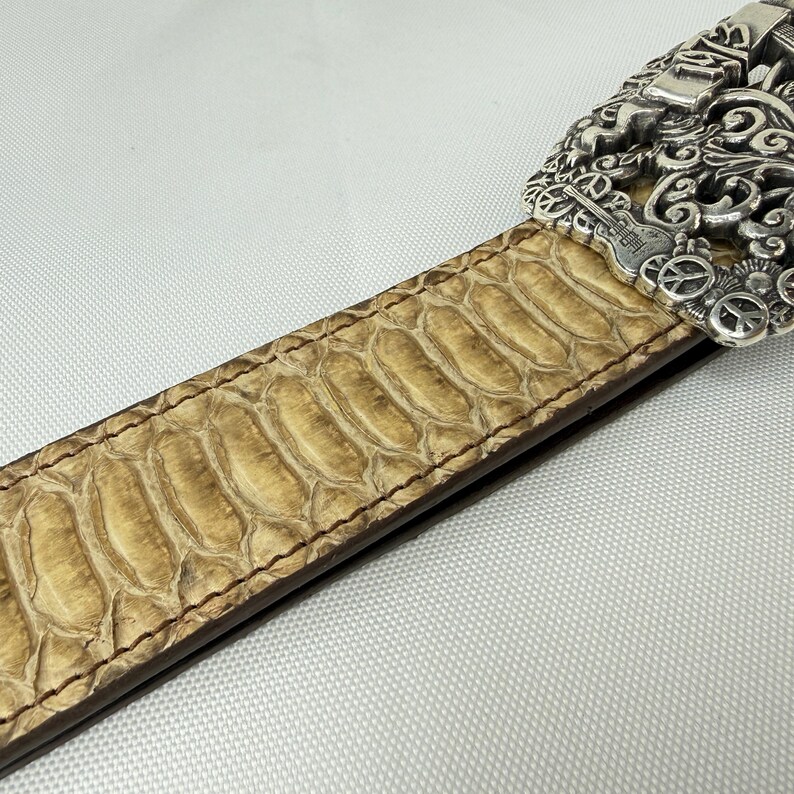 K&ouml;nnte beinhalten: Nahaufnahme eines beigefarbenen Lederg&uuml;rtels mit Schlangenmuster. Der G&uuml;rtel hat eine dekorative Silberschnalle mit aufwendigen Designs. Der G&uuml;rtel ist mit braunem Faden an den Kanten gen&auml;ht.