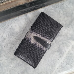 Könnte beinhalten: Eine schwarze Ledergeldbörse mit Schlangenmuster. Die Geldbörse hat eine Klappe mit einem schwarzen Lederriemen und Schnallenverschluss. Die Geldbörse ist rechteckig und scheint geschlossen zu sein.
