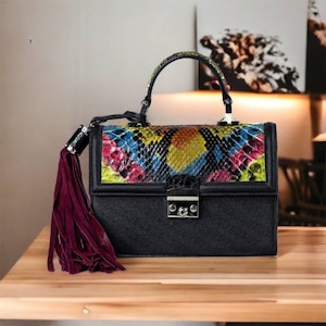 Puede incluir: Un bolso de mano de cuero negro con una solapa de piel de serpiente colorida y un borla de color burdeos. El bolso de mano tiene un asa superior y un cierre de llave.