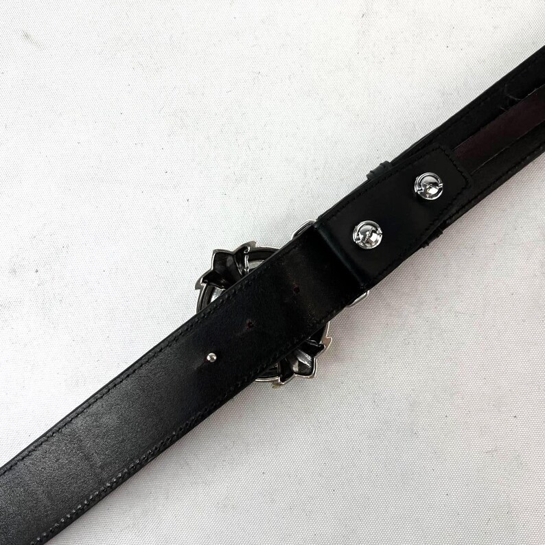 May include: Black leather belt with a decorative silver buckle. The buckle features a floral design. The belt has two silver rivets near the buckle. The belt is angled across a white surface.