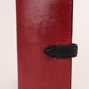 Könnte beinhalten: Ein rotes Lederportemonnaie mit strukturierter Oberfläche und einem Verschluss aus schwarzem Leder. Das Portemonnaie ist rechteckig und vertikal ausgerichtet. Die Nähte sind mit rotem Faden sichtbar. Ein stilvolles Accessoire.