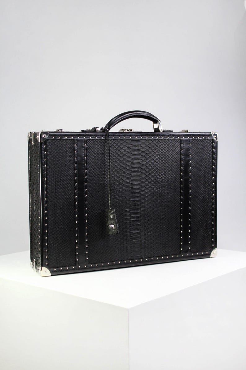 May include: A black rectangular suitcase with a textured snakeskin pattern. The suitcase has a black handle, silver corner protectors, and silver studs along the edges. A small tag hangs from the handle. The suitcase is sitting on a white surface.