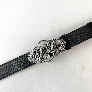 Może przedstawiać: Czarny pasek ze srebrną klamrą. Klamra ma szczegółowy wzór węża przeplatanego liśćmi i jagodami. Pasek ma teksturowaną, wzorzystą powierzchnię.