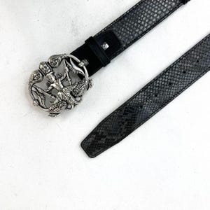 May include: A black belt with a snakeskin pattern and a decorative silver buckle. The buckle features a detailed design with a central figure and ornate embellishments. The belt is made of leather and has a classic design.