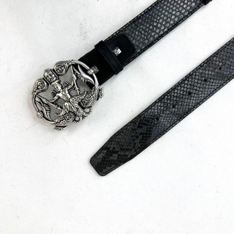 May include: A black belt with a snakeskin pattern and a decorative silver buckle. The buckle features a detailed design with a central figure and ornate embellishments. The belt is made of leather and has a classic design.