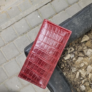 Könnte beinhalten: Ein rechteckiges Portemonnaie in tiefem Rot mit einer glänzenden, geprägten Krokodilleder-Struktur. Das Portemonnaie hat eine schmale, längliche Form mit sichtbaren Nähten an den Kanten und einer glatten, polierten Oberfläche.