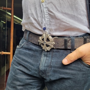 May include: A silver-colored belt buckle in the shape of a cross, adorned with intricate details and a central gemstone. The buckle is attached to a gray snakeskin-patterned belt, worn with blue jeans and a white shirt.