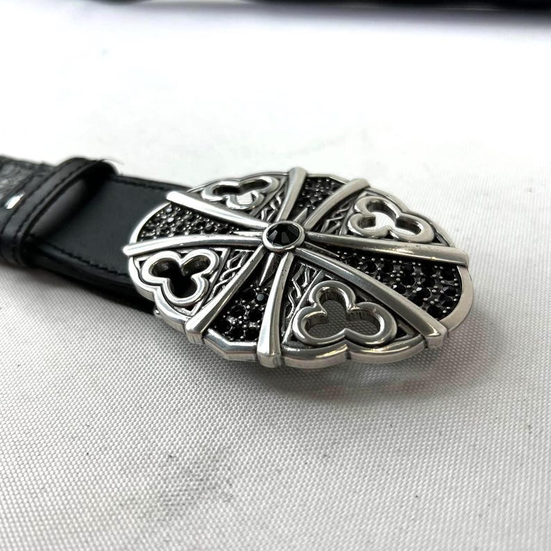 K&ouml;nnte beinhalten: Schwarzer Lederg&uuml;rtel mit silberner Schnalle. Die ovale Schnalle hat einen zentralen schwarzen Stein und ein dekoratives Muster mit schwarzen Steinen. Der G&uuml;rtel liegt auf einer wei&szlig;en Oberfl&auml;che.