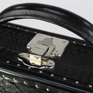 May include: Close-up of a black, textured case with a silver metal clasp and handle. The case has a crocodile skin pattern and is adorned with small silver rivets. The handle is made of black leather.