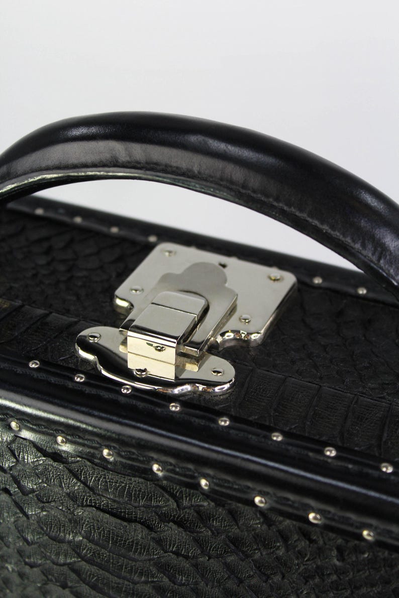 May include: Close-up of a black, textured case with a silver metal clasp and handle. The case has a crocodile skin pattern and is adorned with small silver rivets. The handle is made of black leather.