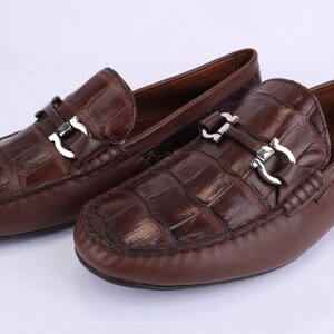 May include: A pair of brown leather loafers with a crocodile pattern. Each shoe features a silver-colored buckle and a rounded toe. The shoes are displayed on a white surface.