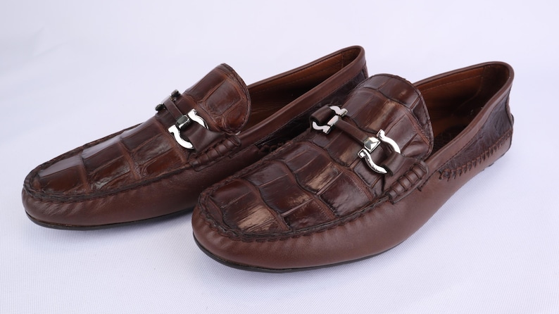 May include: A pair of brown leather loafers with a crocodile pattern. Each shoe features a silver-colored buckle and a rounded toe. The shoes are displayed on a white surface.