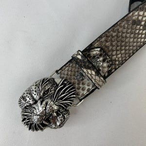Pode incluir: Um cinto com uma fivela prateada detalhada em forma de cabe&ccedil;a de le&atilde;o. A correia apresenta um padr&atilde;o de pele de cobra em tons de cinza e preto. A fivela &eacute; intrincadamente projetada, mostrando o rosto do le&atilde;o.