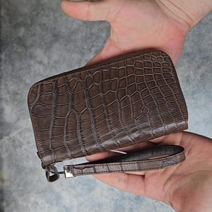 May include: A brown, rectangular wallet with a crocodile skin pattern. The wallet has a zip closure and a wrist strap. The wrist strap is attached to the wallet with a silver-coloured metal clasp.