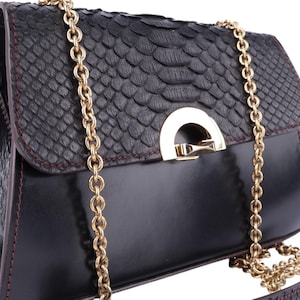 Peut inclure: Sac à main en cuir noir avec rabat texturé et chaîne dorée. Le sac est doté d'un fermoir doré et de coutures rouges contrastantes. Le design intègre un motif peau de serpent sur le rabat.