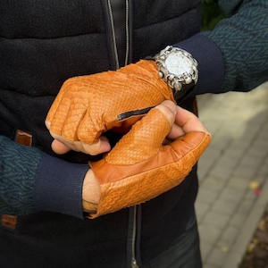 Peut inclure: Gants en cuir orange avec une texture peau de serpent et une fermeture éclair noire. Les gants sont sans doigts. Une montre argentée avec une lunette décorative est portée au poignet.