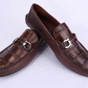 May include: A pair of brown leather loafers with a crocodile pattern. The shoes have silver-colored buckles and are displayed on a white background. These driving shoes are a classic style.